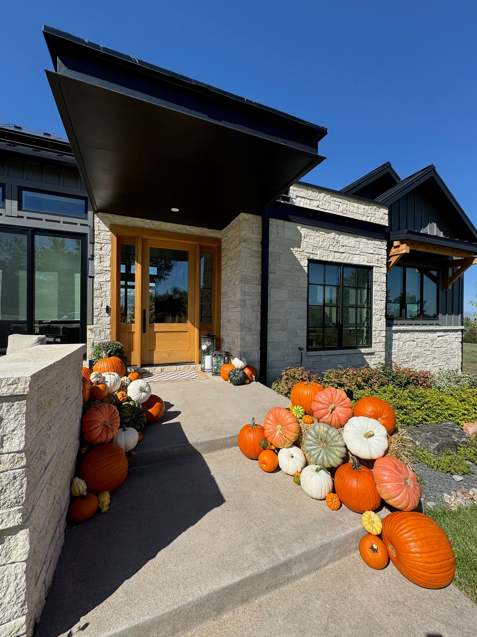 A modern stacked-stone house with a light wood door and black overhang, with pumpkin clusters on both sides of the walkway